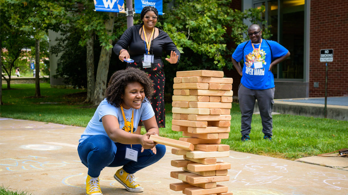 A Webster student carefully removes a block from a lawn-sized game of falling tower while other students look on. 