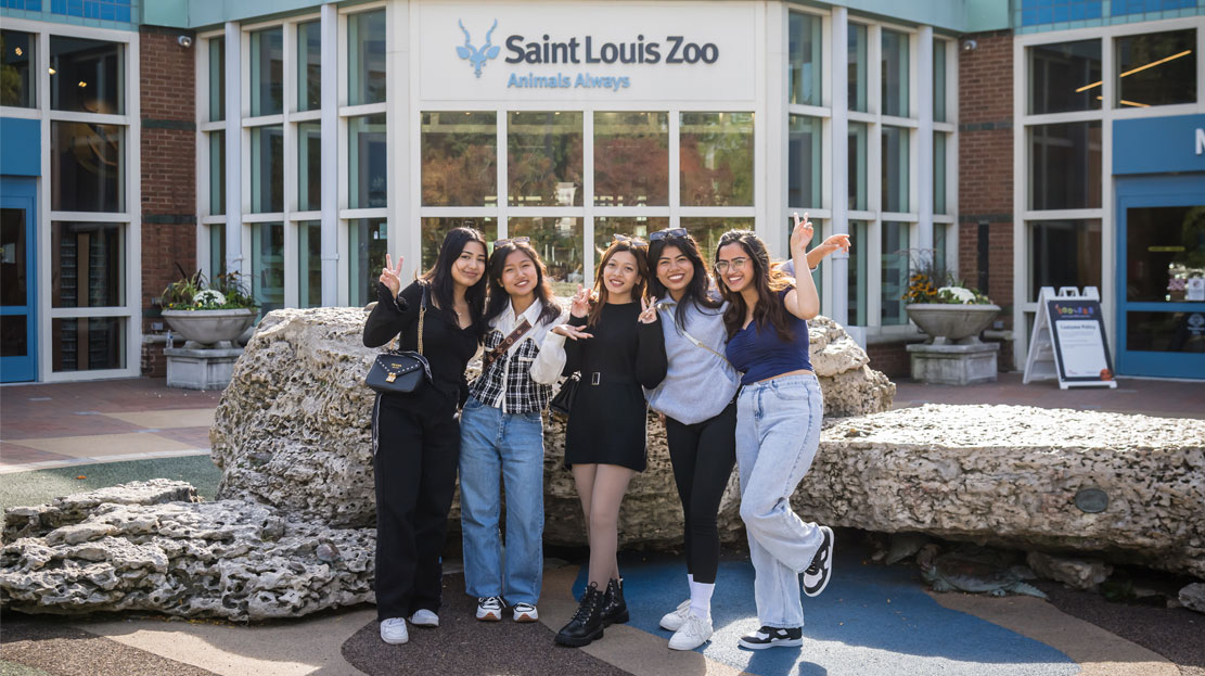 A group of Webster students pose and smile arm in arm outside the St. Louis Zoo entrance.