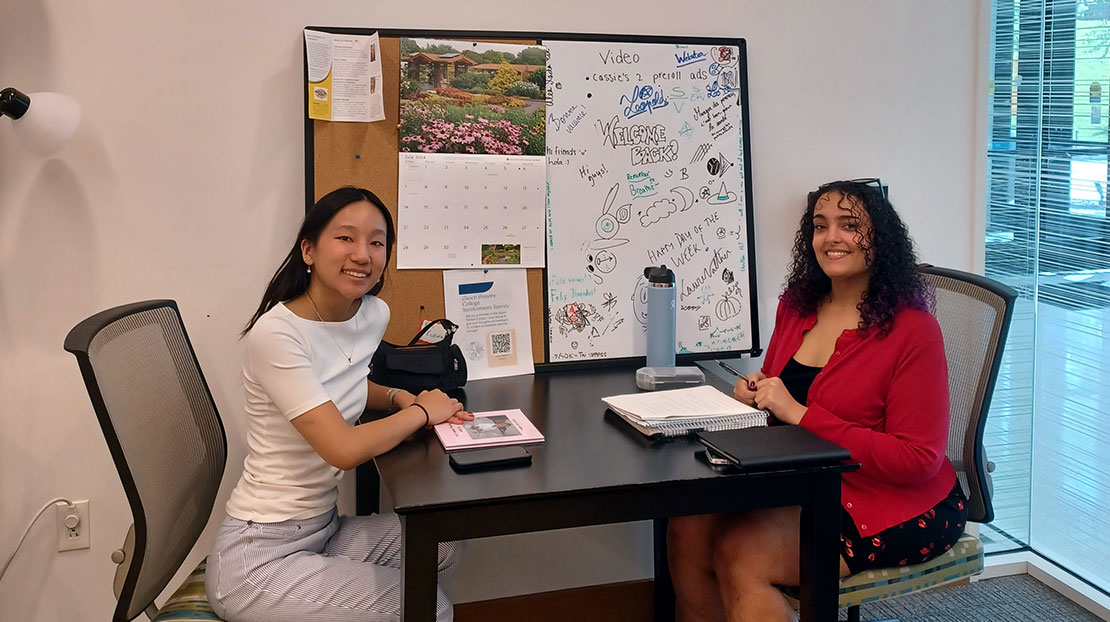 Two students site across from each other at a table with paper and books sitting on it.