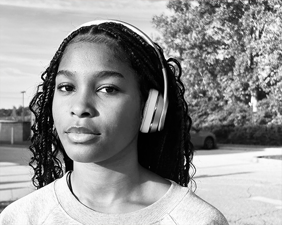 Black and white portrait of a young woman with braids wearing headphones and a sweatshirt looking out of the image with her head slightly angled.