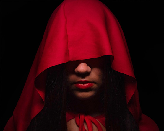 Portrait of a red cloak that drapes over and covers the eyes, leaving the nose and red lips showing. The ties of the cloak are visible around the neck.