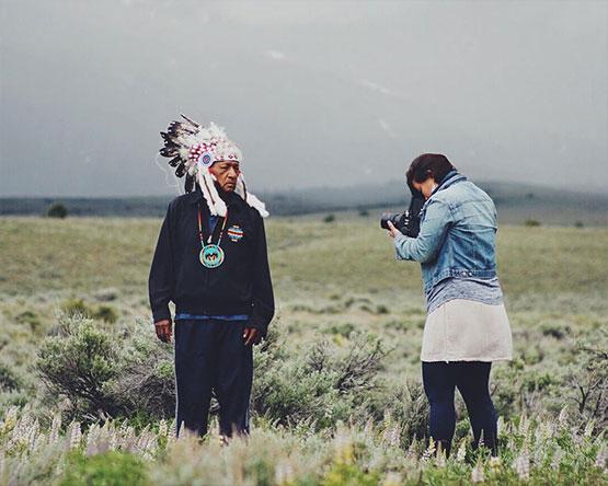 Man wearing native feathered headdress stands in front of Matika Wilbur who is taking his photo, both standing in a field of sage brush.