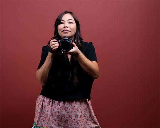 Matika Wilbur, self-portrait, stands against a red-toned background holding a camera up near her face.