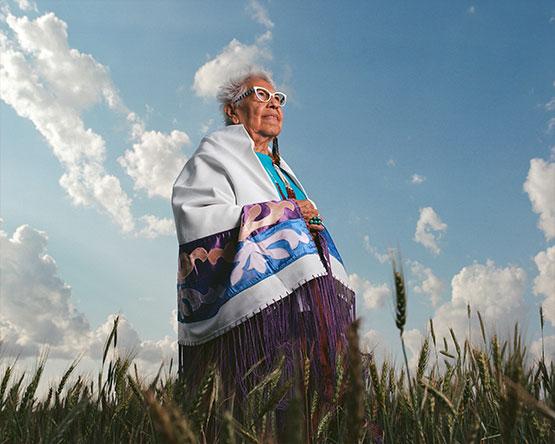 Elder native woman with white glasses looks skyward while standing in wheat field wearing white shawl with purple trim and blue and purple decorated stripes