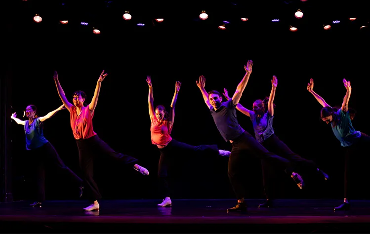 Several dancers stand in a line and raise their hands in the air on a darkly lit stage.