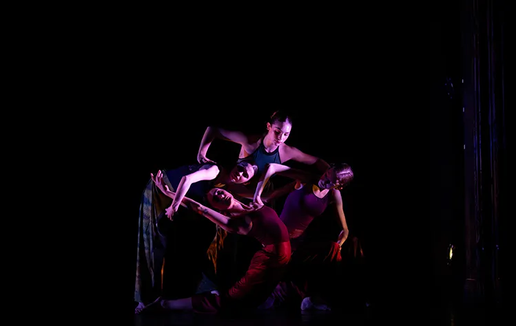 Four dancers create a circle on a darkly lit stage.