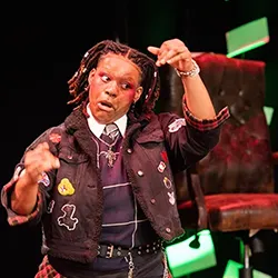 Webster Conservatory actor with locs and dramatic eye makeup gestures dynamically on stage in a patch-adorned jacket in front of a leather armchair.
