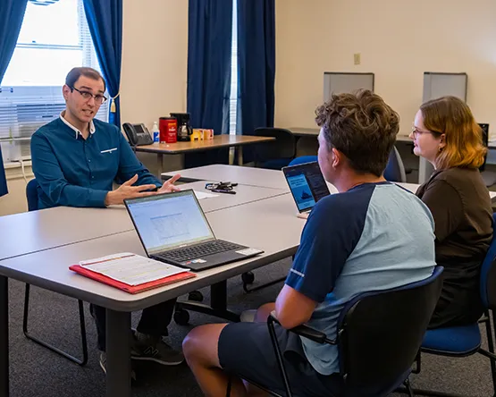 Dr. Dani Belo sits with two research assistants at a table with laptops, talking animatedly