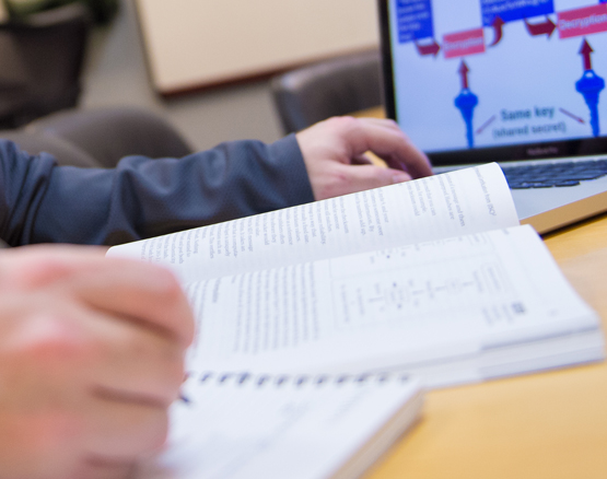 hands of student with an open textbook beside a laptop with cybersecurity information on the screen.